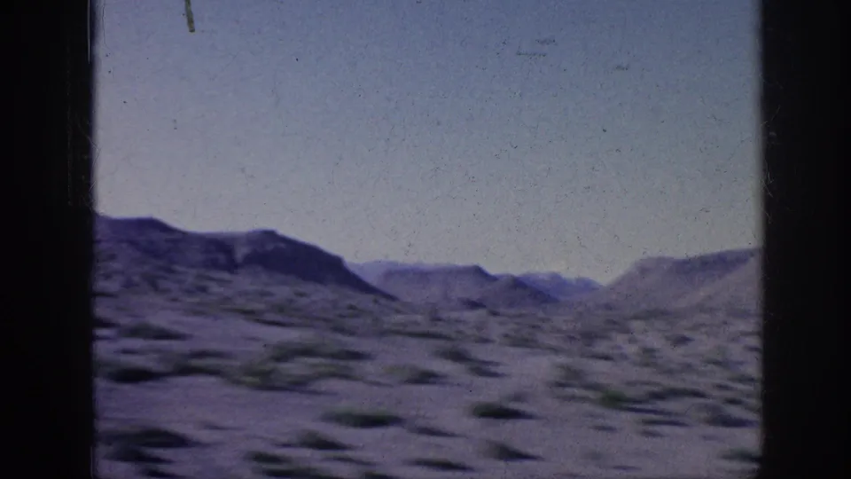 1963 8mm home-movie frame showing blurred scrub and sandy ground in the foreground with low hills and mesas in the distance.