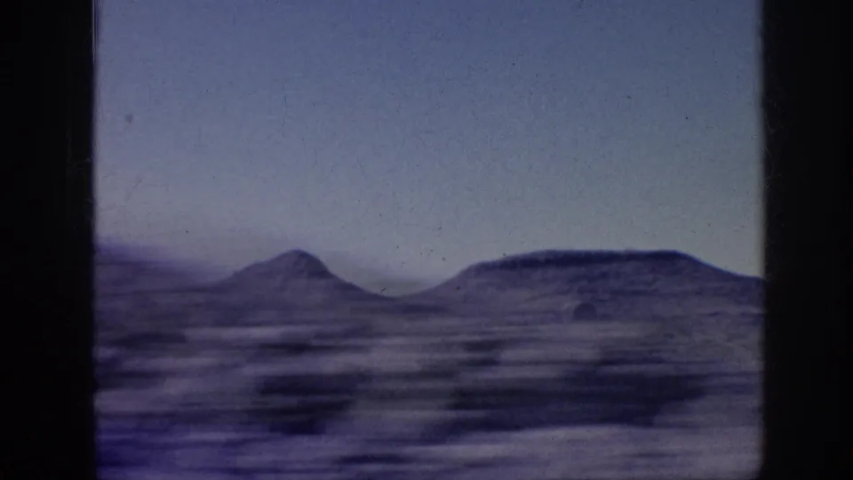 1963 8mm frame of a motion-blurred desert plain, scattered scrub, and rounded distant hills beneath an open pale sky.