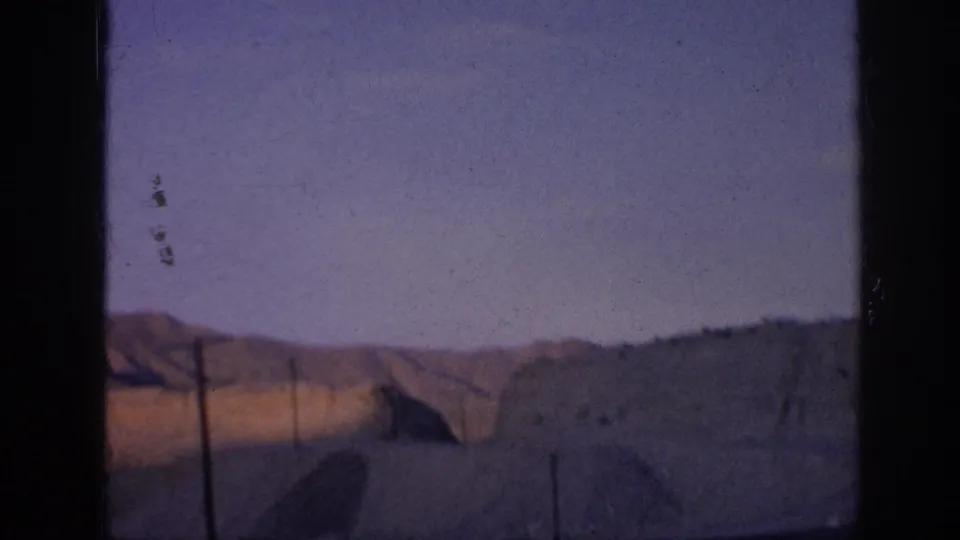 8mm frame showing rolling hills and roadside posts beneath open sky, with dust marks and dark scan borders visible.