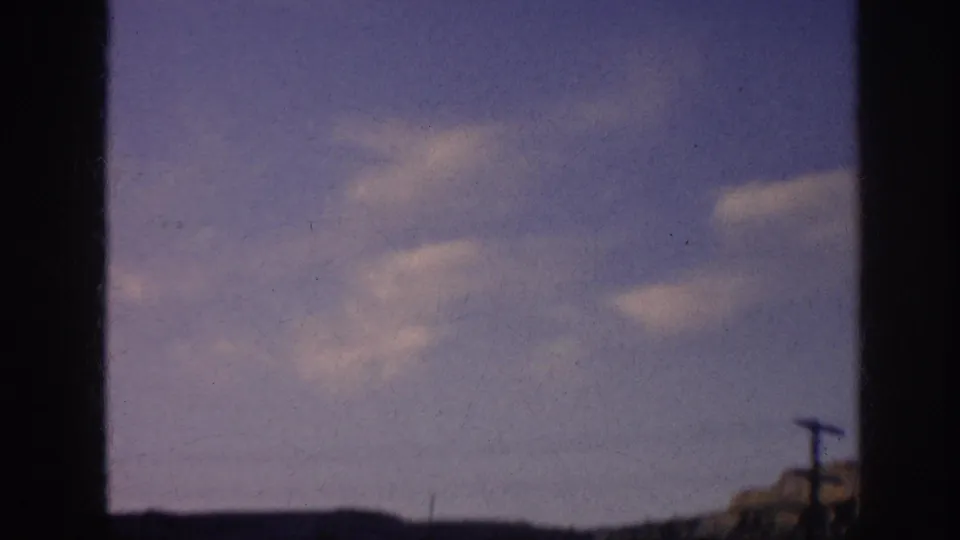 1963 8mm frame with low terrain, a utility pole, overhead wire, and clouds under a blue sky.