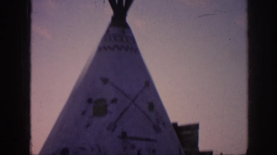 1963 8mm frame showing a teepee-like structure with painted motifs beside a building edge under twilight-colored sky.