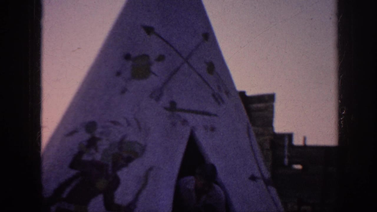 1963 Memphis home-movie frame showing a painted teepee-style structure with figures, arrows, wooden buildings, grass, and a wagon wheel nearby.