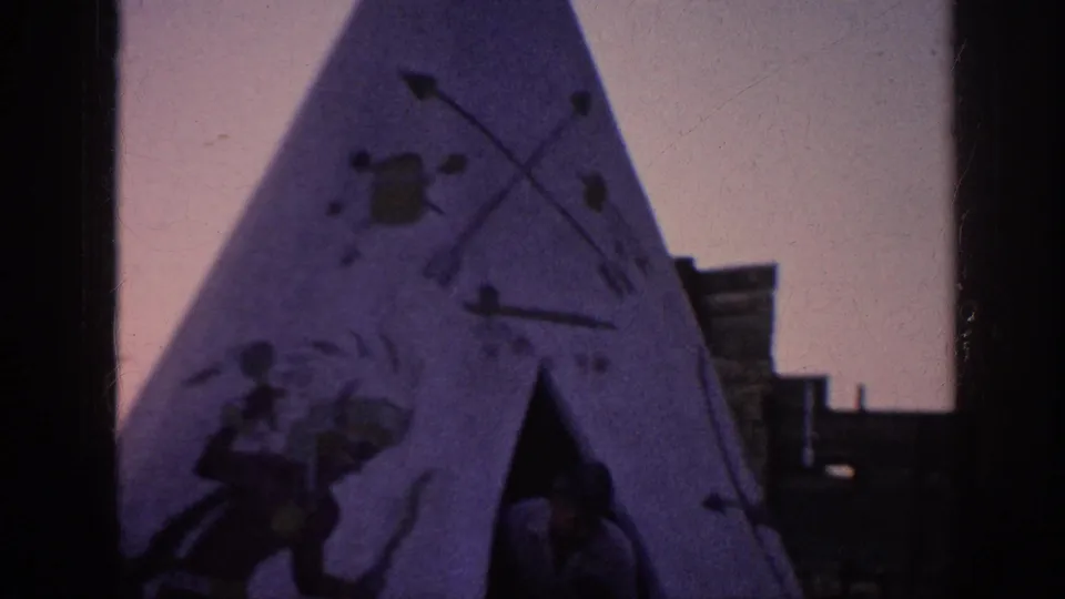 1963 Memphis home-movie frame showing a painted teepee-style structure with figures, arrows, wooden buildings, grass, and a wagon wheel nearby.
