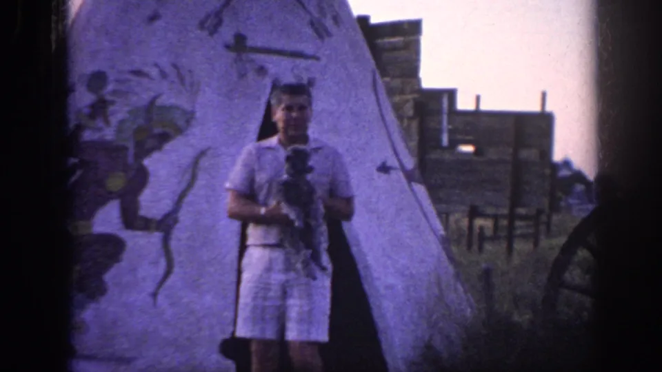 1963 Memphis home-movie frame showing an adult man facing the camera with a small dog in front of a painted teepee and wooden buildings.