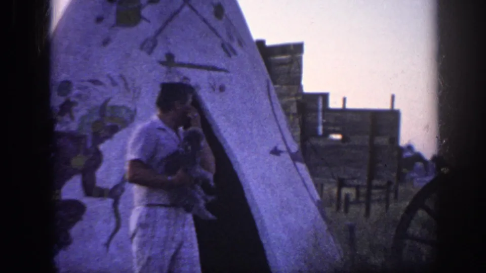 1963 Memphis home-movie frame of a man turned slightly toward a small dog beside the entrance of a painted teepee-style display.