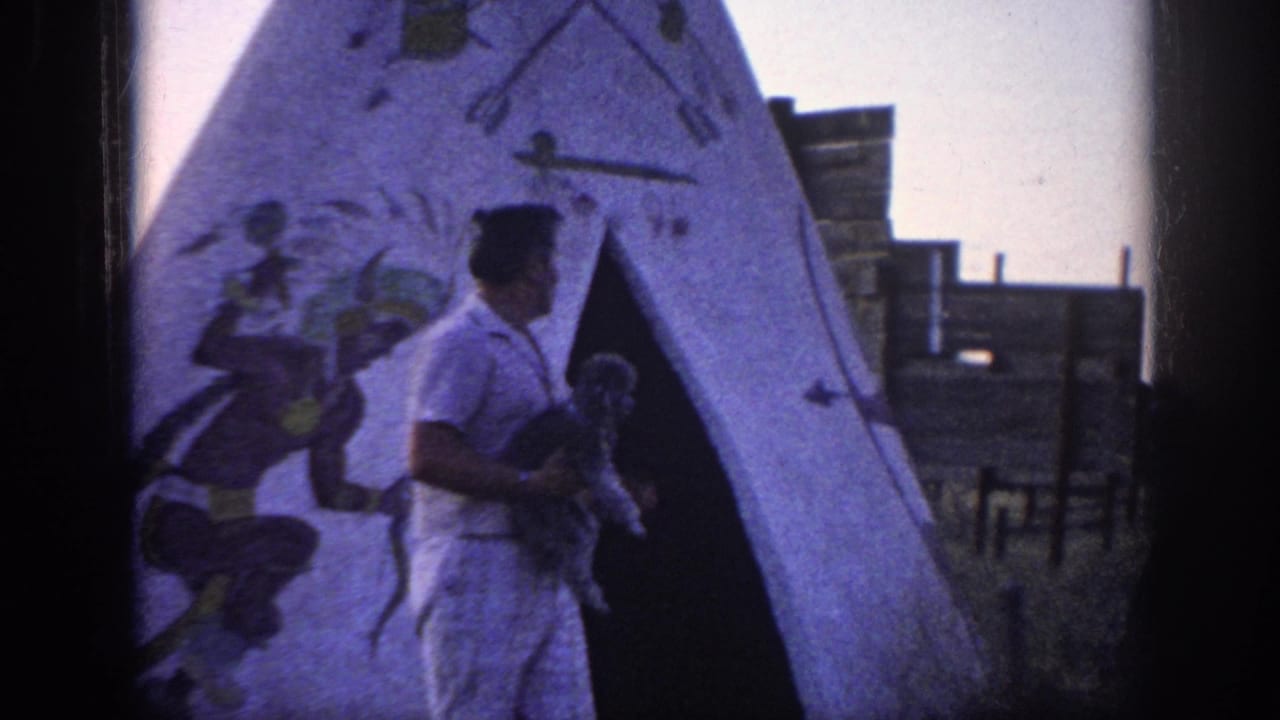 1963 Memphis-area home-movie frame of a man in light clothing holding a poodle near a white teepee with painted figures and arrows.