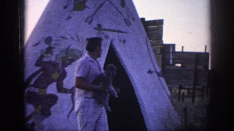 1963 Memphis-area home-movie frame of a man in light clothing holding a poodle near a white teepee with painted figures and arrows.