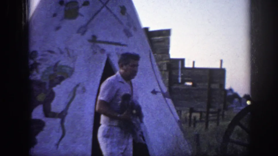 The man holds the poodle while standing closer to the teepee entrance, with rustic structures visible behind him.