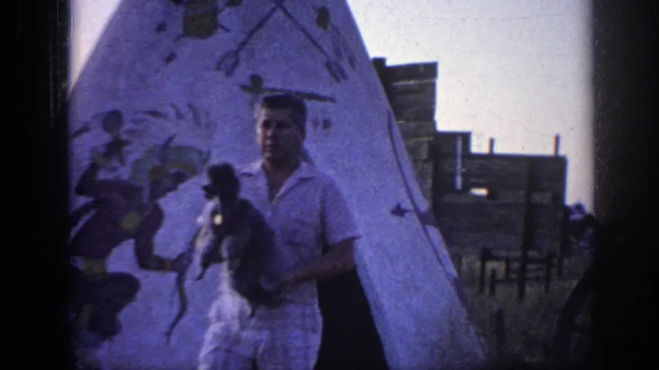 The man faces toward the camera with the poodle in his arms, framed by painted figures on the teepee and wooden structures beyond.