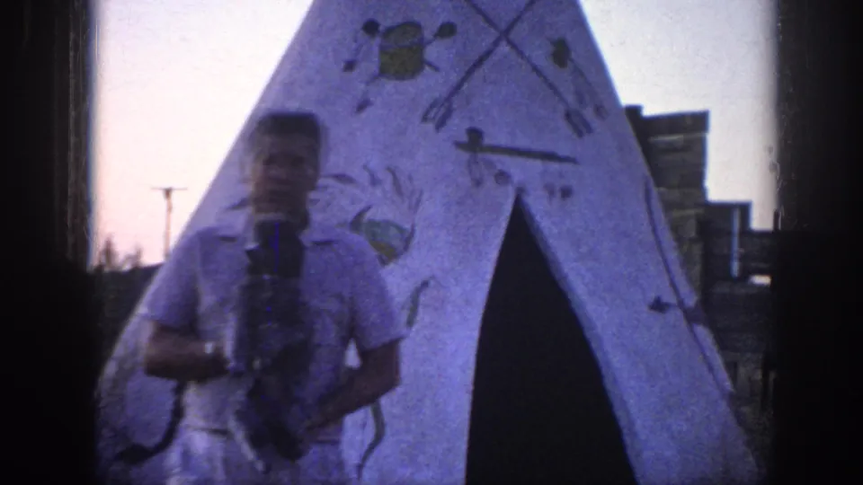 A soft 8mm frame shows the man holding the poodle directly before the teepee opening, with painted arrows and figures around him.