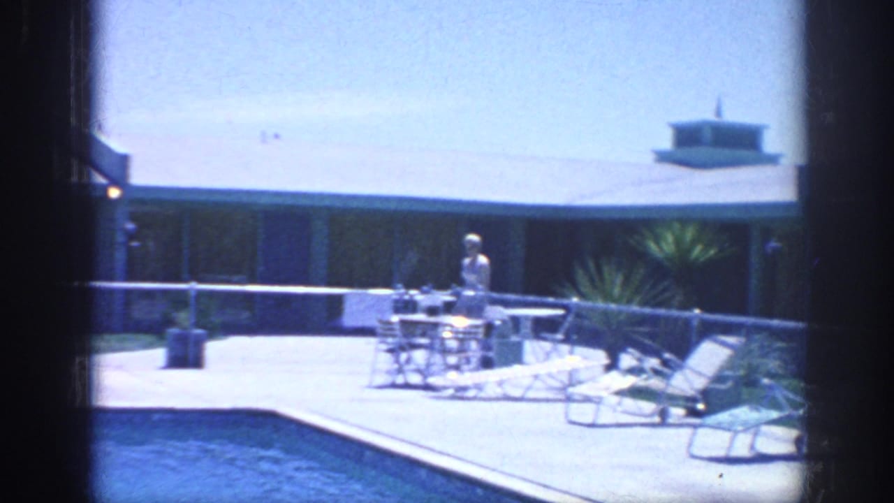 Adult standing near a table and lounge chairs beside a swimming pool.