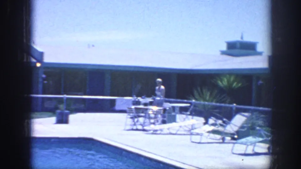 1963 Memphis vacation frame showing an adult by poolside chairs and a table outside a low modern building.