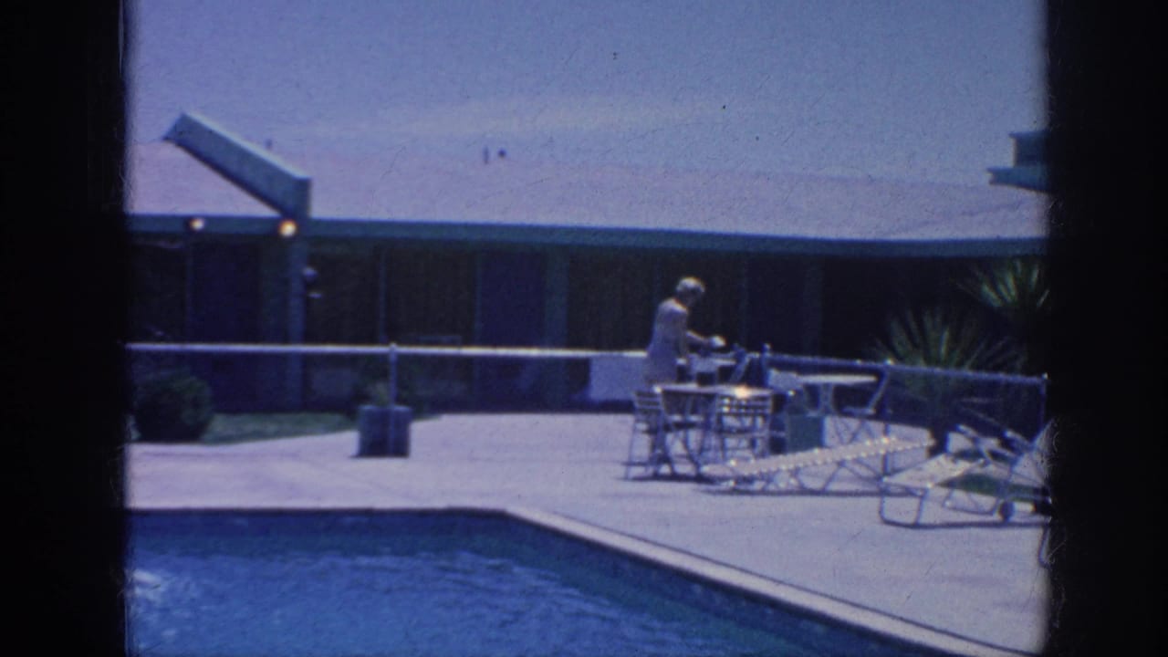 1963 Memphis home-movie frame showing a woman at a sunny poolside patio with chairs, lounge furniture, railing, and a low modernist building.