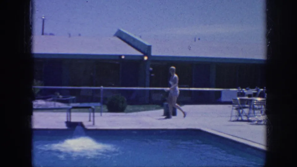 1963 Memphis poolside frame with a woman walking near the pool edge while a small fountain bubbles in the water and patio furniture sits nearby.