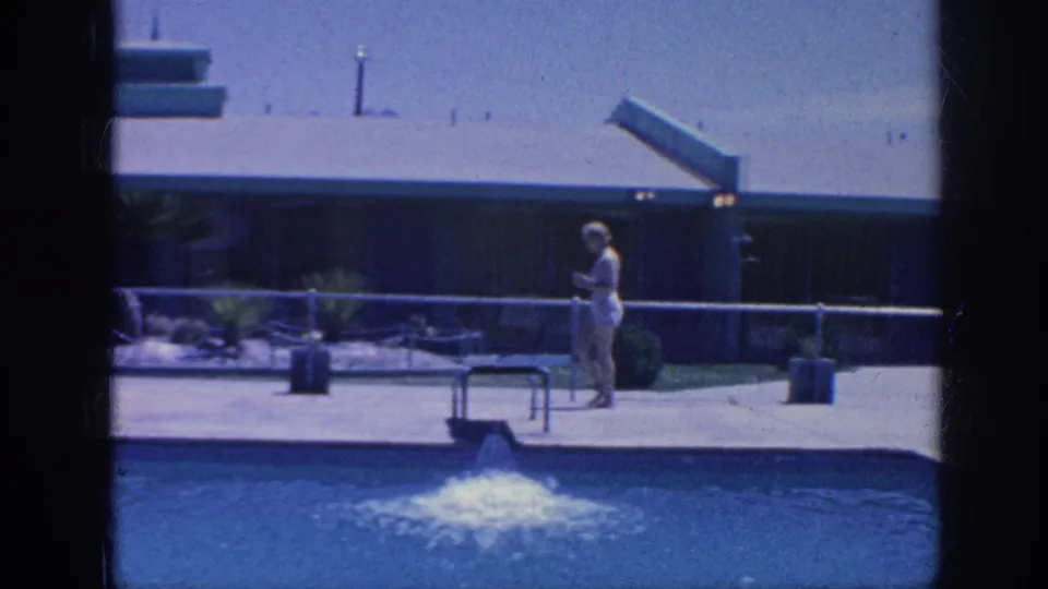 A woman walks beside a Memphis swimming pool, with fountain spray, a metal railing, a low building, and sparse landscaping in the background.