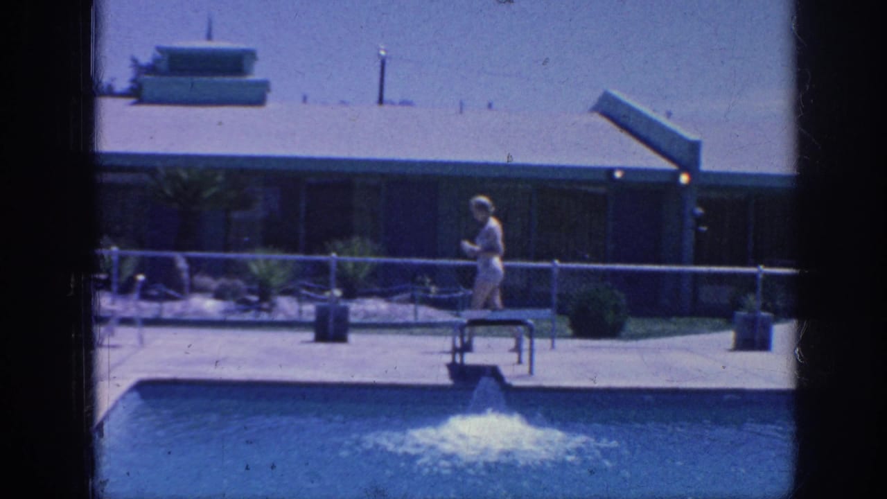 Woman in a light swimsuit walks beside a motel swimming pool with a fountain and low building behind her.
