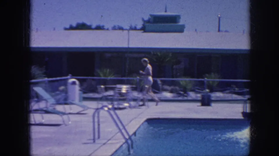 1963 Memphis poolside frame showing the woman near lounge chairs, trash bin, pool ladder rails, planted beds, and the motel building.