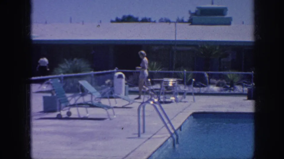 1963 Memphis home-movie frame with a woman near the pool deck, lounge chairs, handrails, trash bin, and a distant person by the building.