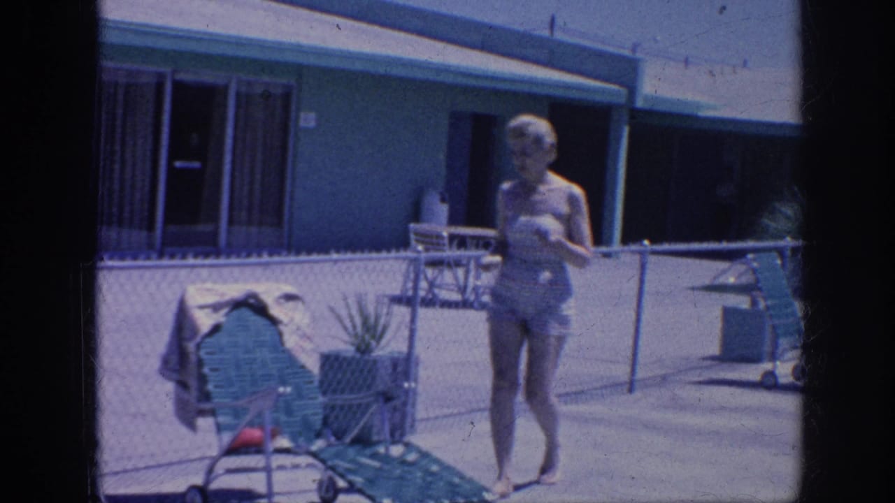 1963 Memphis home-movie frame showing a woman in a patterned swimsuit walking toward a green lounge chair beside a chain-link pool fence.