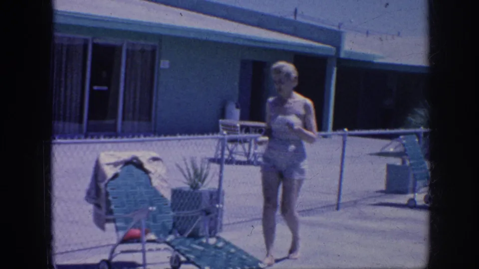 1963 Memphis home-movie frame showing a woman in a patterned swimsuit walking toward a green lounge chair beside a chain-link pool fence.