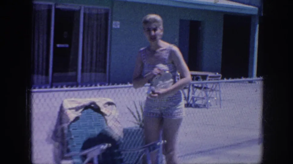 1963 Memphis home-movie frame with a woman in a patterned swimsuit standing by the lounge chair and holding a purse or small bag.