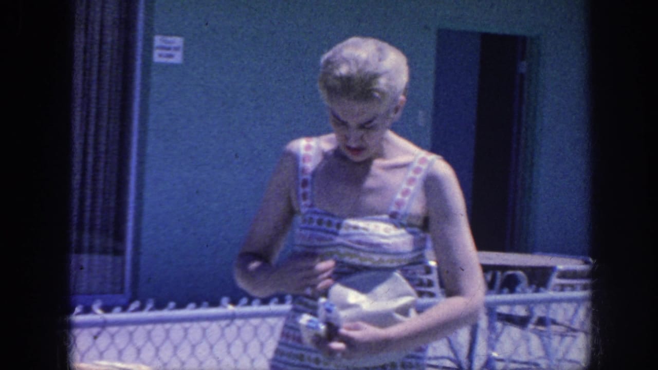 In Memphis 1963 home-movie footage, an adult woman in striped summer clothing looks down at a small object near a poolside fence.