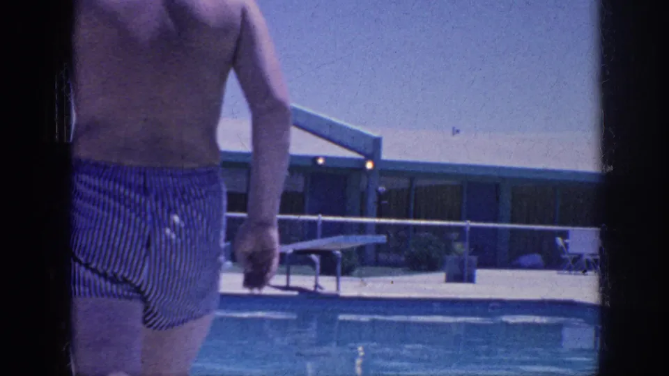 An adult man in striped swim trunks walks through the Memphis 1963 pool area, with the pool, fence, and diving board behind him.