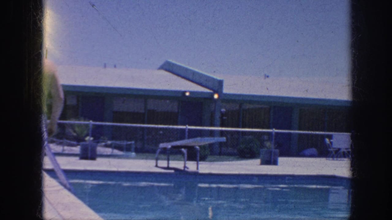 Poolside view with a person partly visible, diving board, fence, and low building.
