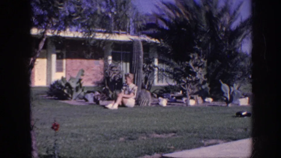 1963 Memphis home-movie frame showing a woman relaxing on a grassy yard before a brick house with cacti and tropical plants.