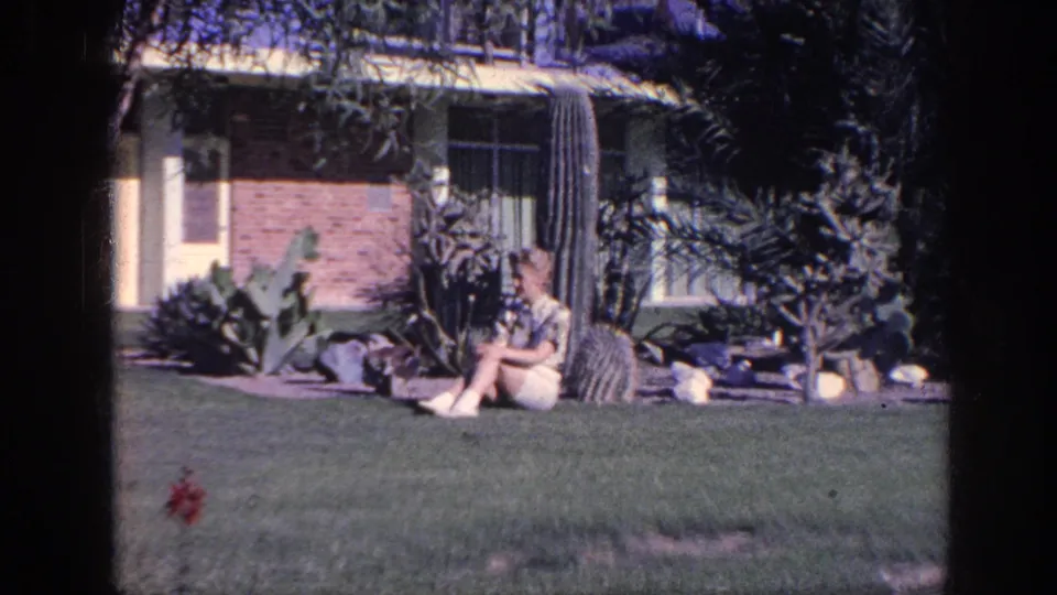 1963 Memphis yard scene with a woman seated on the grass outside a brick house, framed by cacti, shrubs, and trees.
