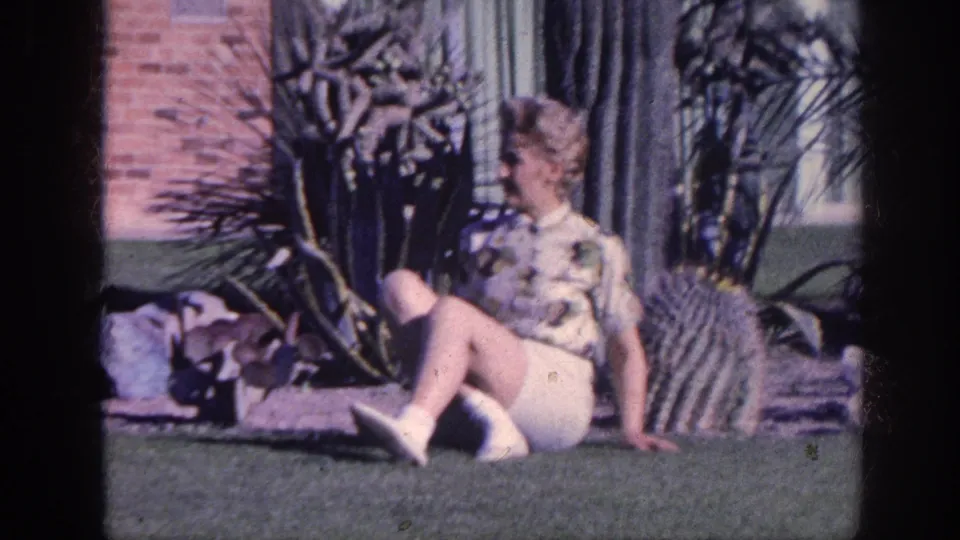 The final frame shows the woman shifting her seated pose on the grass beside tall cacti outside the brick residence.