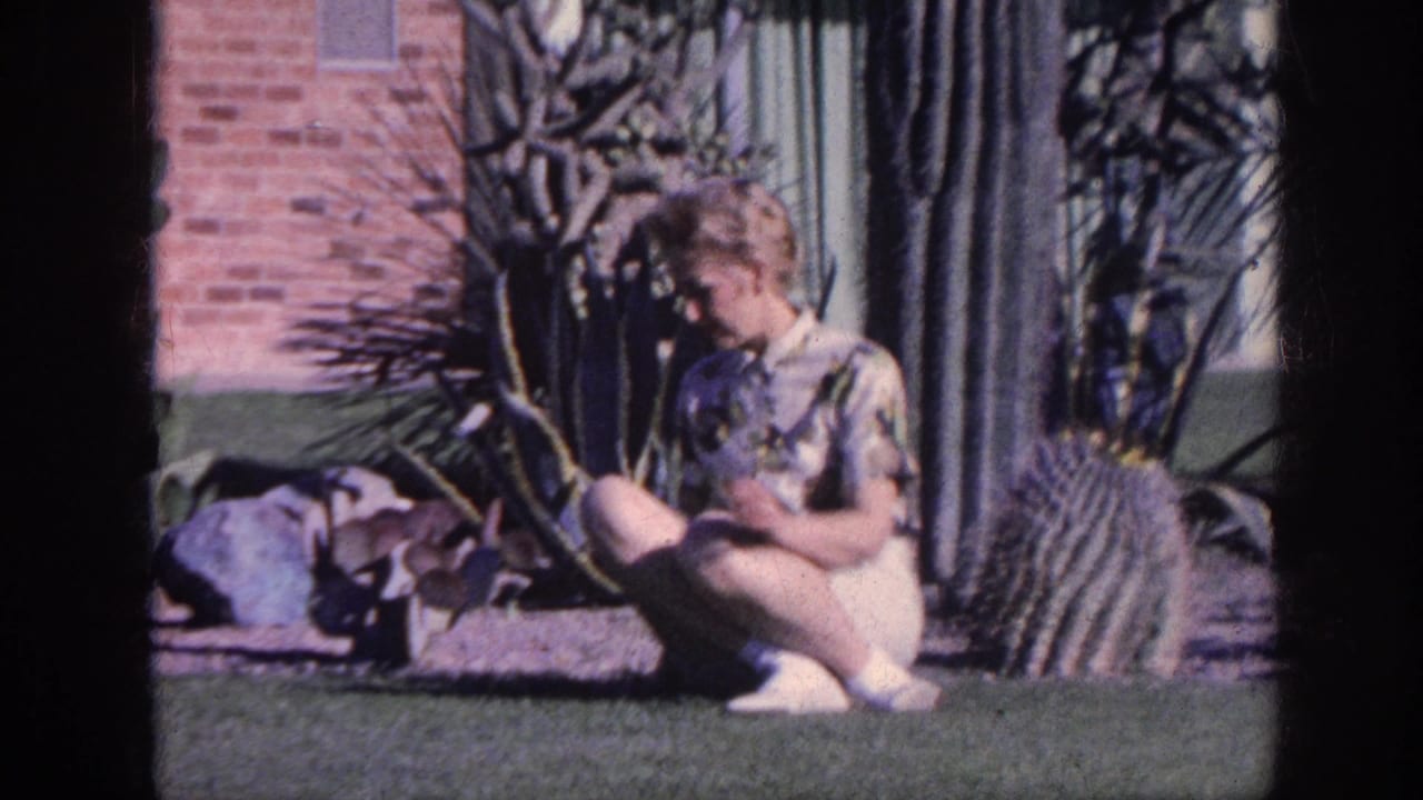 In 1963 Memphis home-movie footage, a woman in a patterned shirt and white shorts crouches by cactus plants in a sunlit yard.