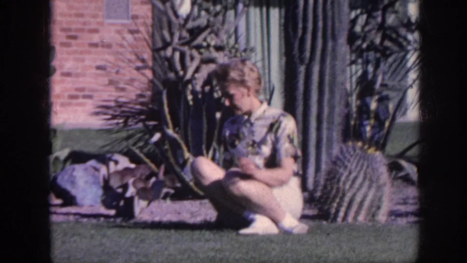 In 1963 Memphis home-movie footage, a woman in a patterned shirt and white shorts crouches by cactus plants in a sunlit yard.