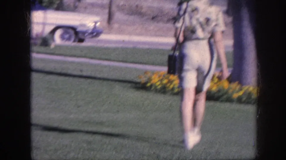 The woman walks away from the camera across a Memphis lawn, with a flower bed, sidewalk, and classic car visible behind her.