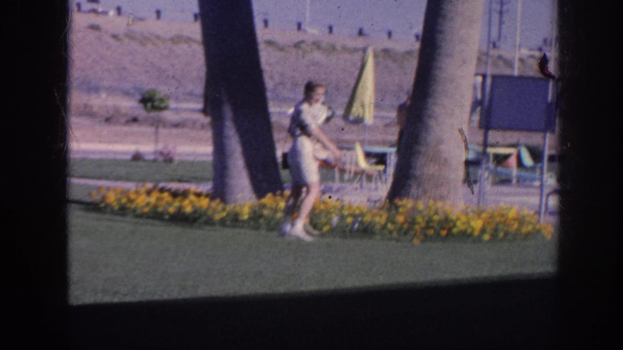 In a 1963 Memphis home-movie frame, a woman in a light dress stands by a flower bed between palm trunks with patio furniture behind her.