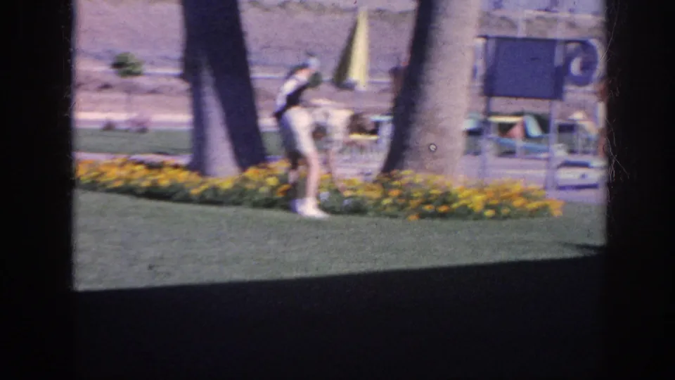 The Memphis 1963 home-movie frame shows a woman leaning toward yellow flowers as another adult stands nearby among palm trees and patio chairs.