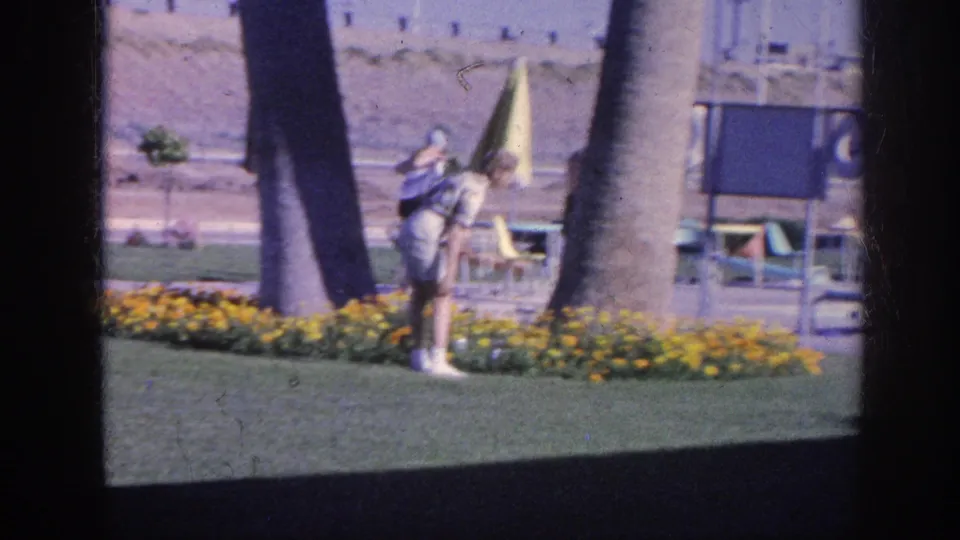 A 1963 Memphis home-movie frame shows two adults near a landscaped flower bed, with palm trunks, lawn, patio seating, and a yellow umbrella visible.