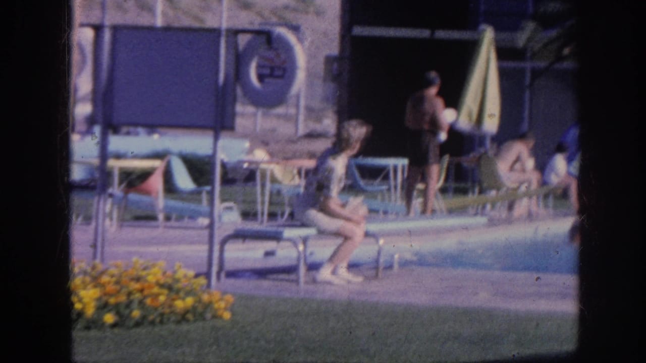 Woman seated beside a motel pool while other guests stand near lounge chairs and an umbrella.