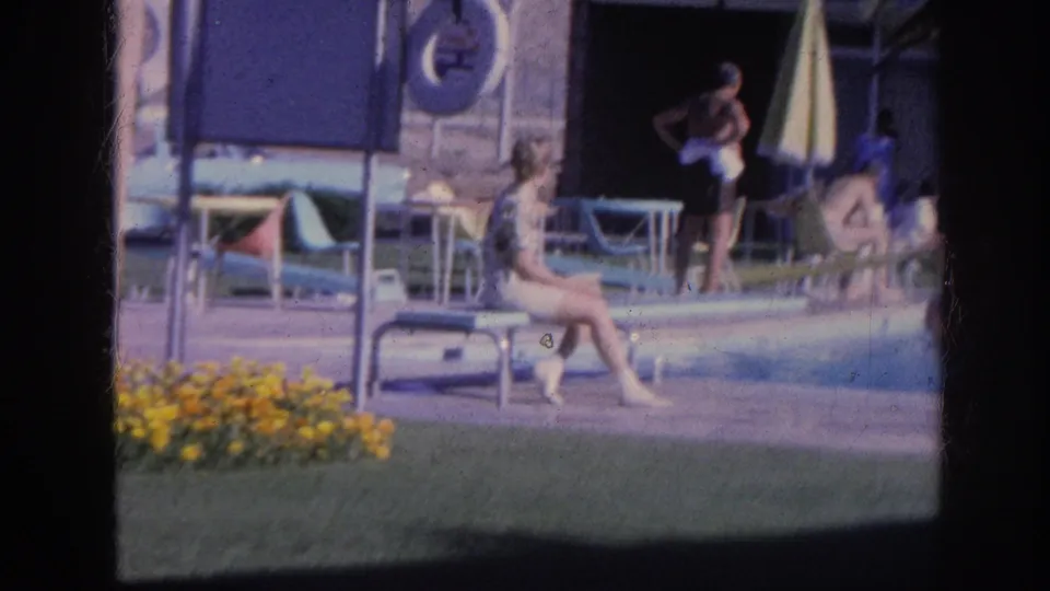 1963 Memphis Holiday Inn poolside frame with a woman seated near the water, guests behind her, lounge chairs, flowers, and motel exterior details.