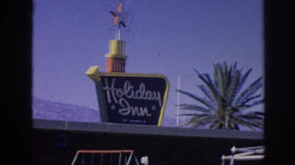 1963 Memphis, Tennessee 8mm frame showing the Holiday Inn sign from a similar visitor angle, with the sign tower centered, palm fronds at right, and blue sky behind.