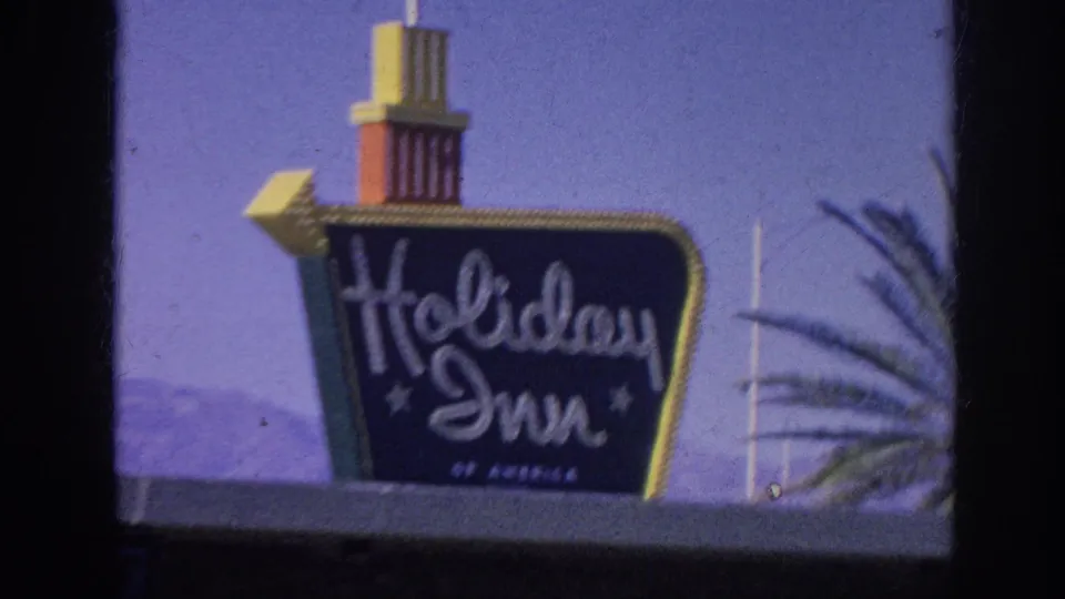 1963 Memphis, Tennessee frame with a closer handheld view of the Holiday Inn sign, showing the tower top, cursive brand lettering, blue sky, and partial palm tree.