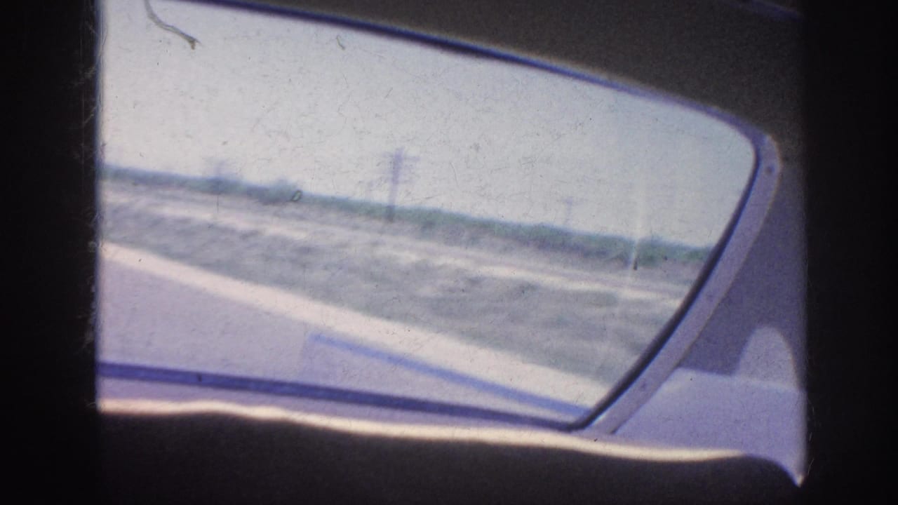 View through a car side window toward a paved road and flat roadside landscape.