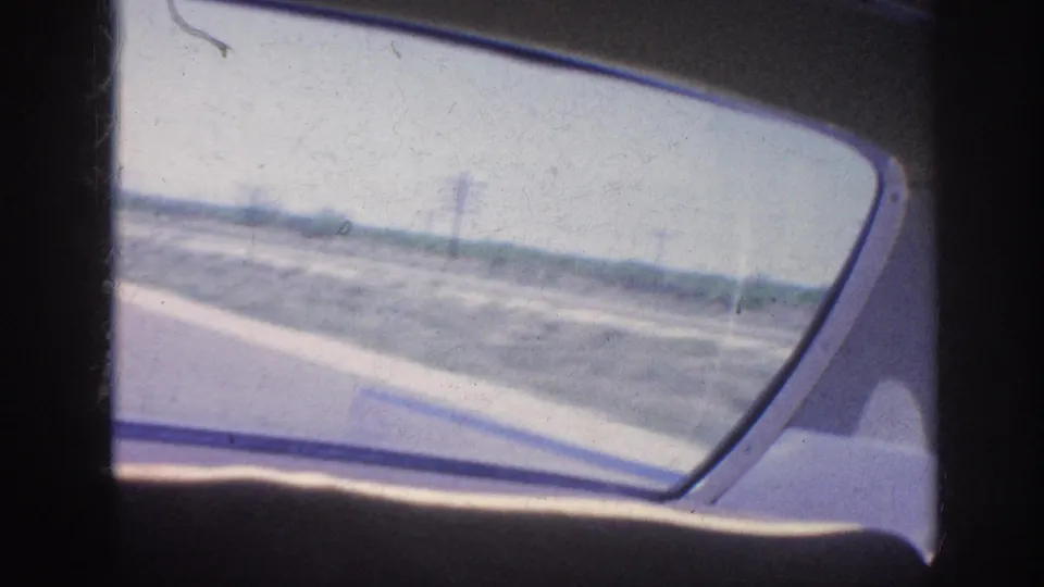 1963 Memphis-area 8mm frame looking from inside a moving car through the side window at road, shoulder, and open flat terrain.