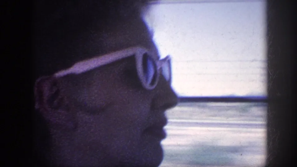 In the supplied 1963 Memphis home-movie clip, a woman wearing white-framed sunglasses is shown in profile against the bright passing roadside.