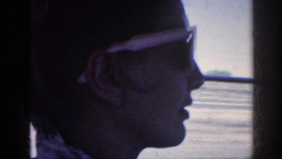 1963 8mm home-movie frame of a car passenger in white sunglasses, partly silhouetted against the passing view.