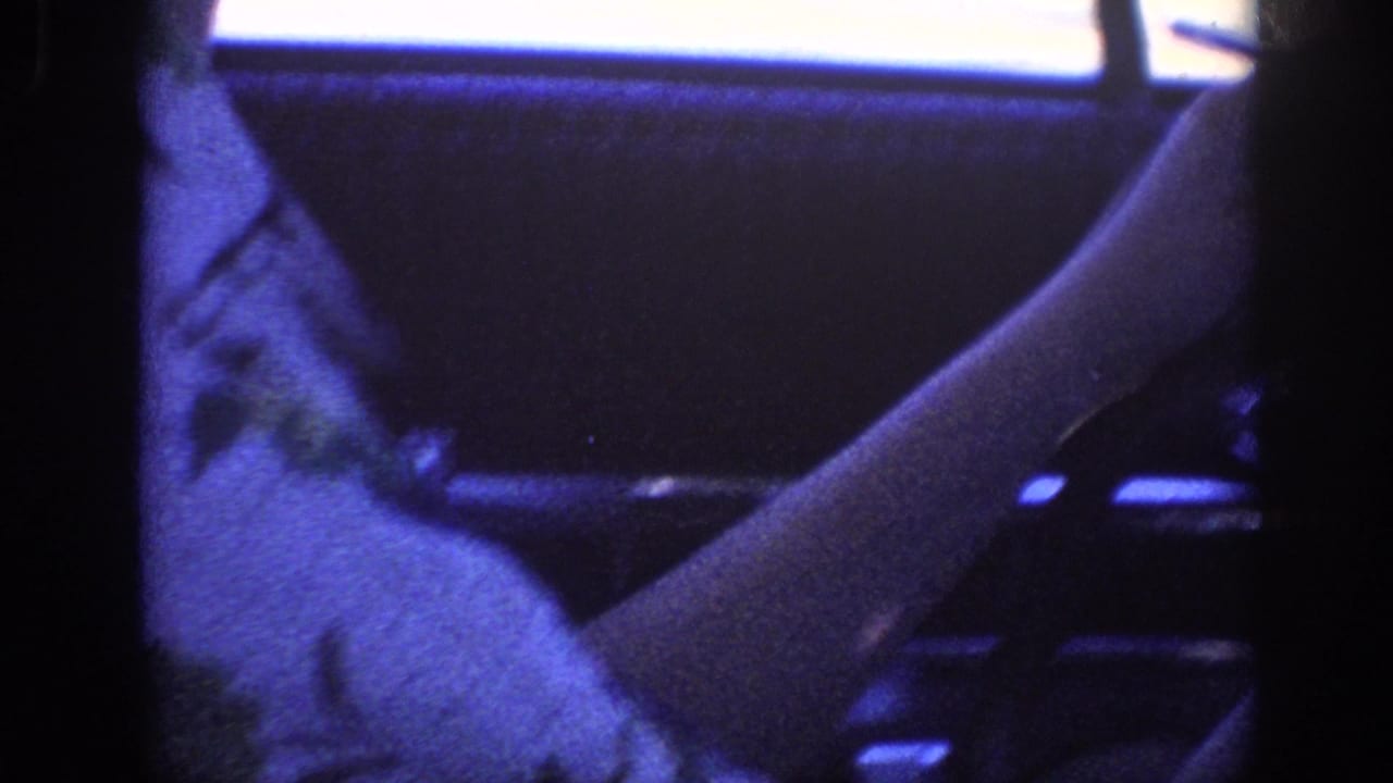 Dark car interior close-up with arm visible near steering column area, heavy blue-purple color cast, 8mm film grain.
