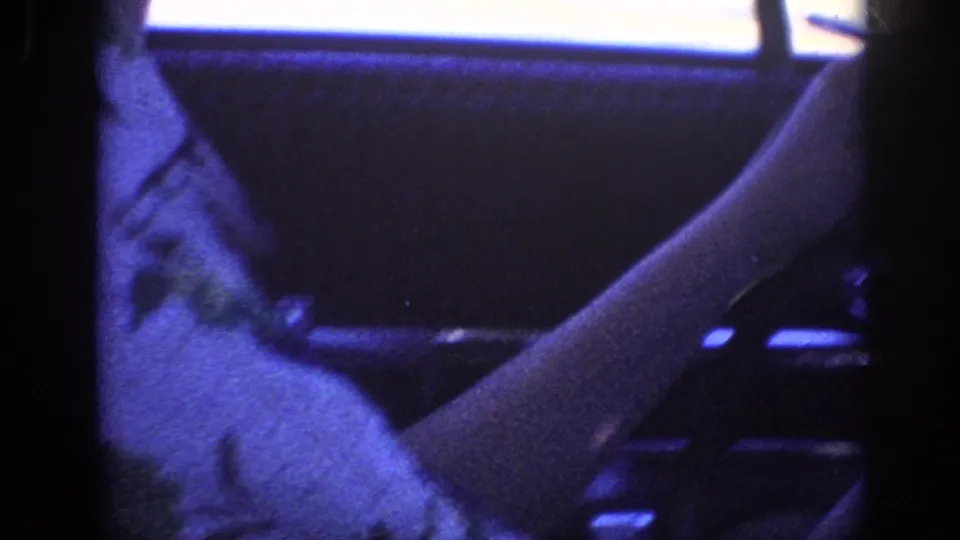 Dark interior detail of a 1960s car, arm or hand visible near the steering column, strong blue-purple color cast and film grain from 8mm stock.