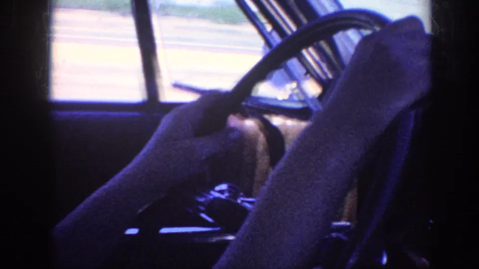 Driver's hands on a large-diameter steering wheel with blurred open highway visible through the side window, filmed on 8mm in a 1960s American automobile, Memphis Tennessee 1963.