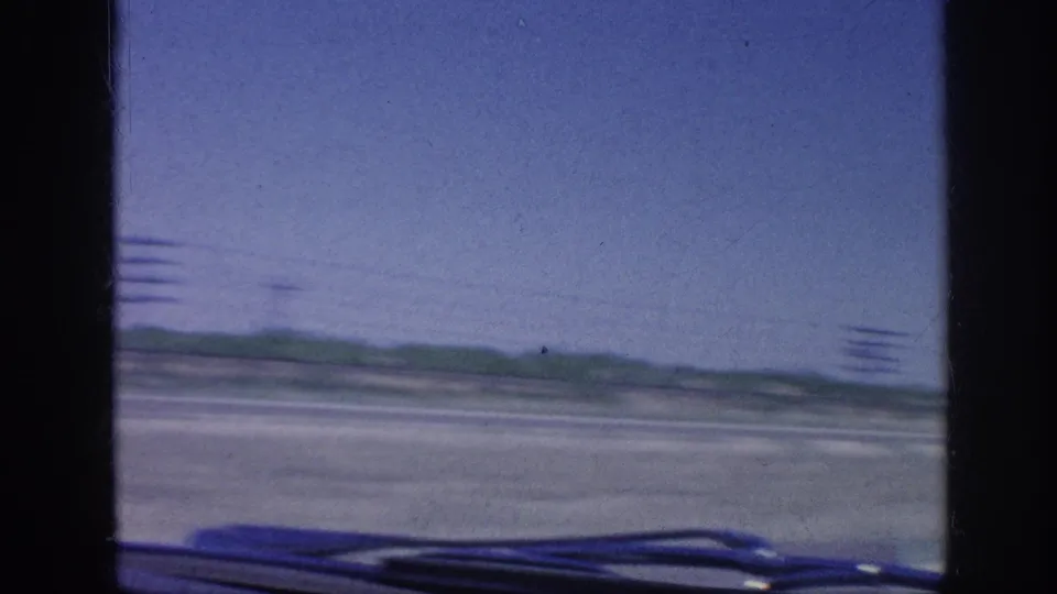 1963 8mm driving footage frame showing the view through a car windshield across open rural roadside terrain, with power poles and the hood edge in the foreground.