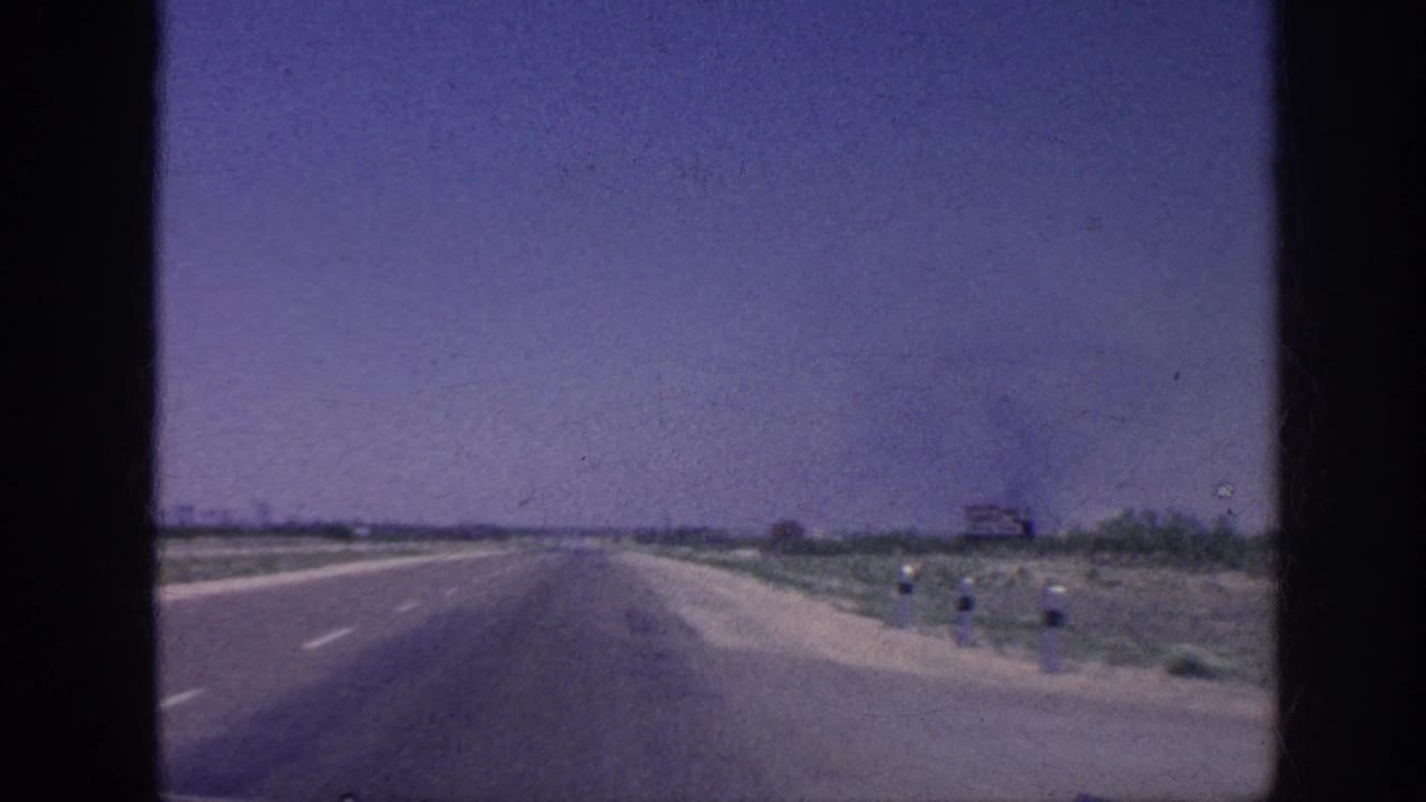 8mm windshield POV of a straight American highway, 1963, with a dark smoke plume on the left horizon, sparse roadside scrub, and a distant billboard at right.